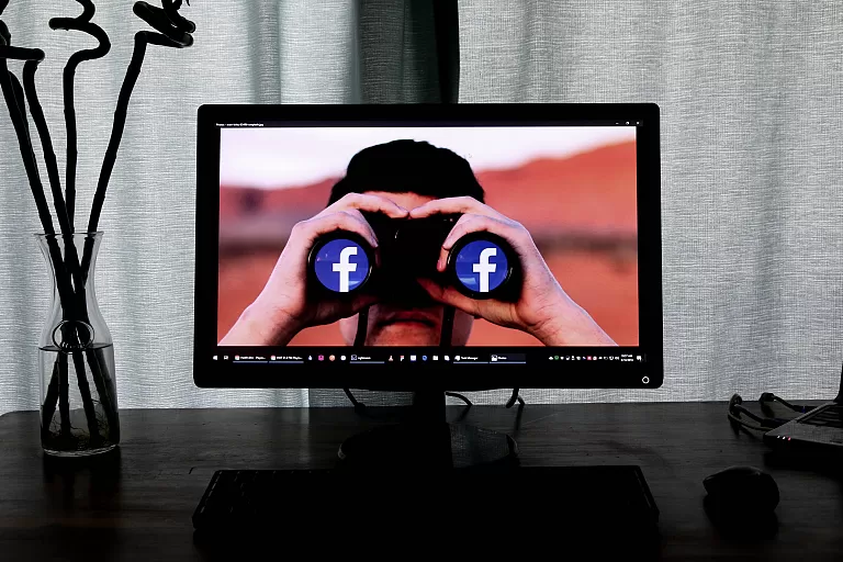 A computer on a desk. The computer shows a picture of someone holding binoculars; the Facebook logo is reflected in the binoculars.