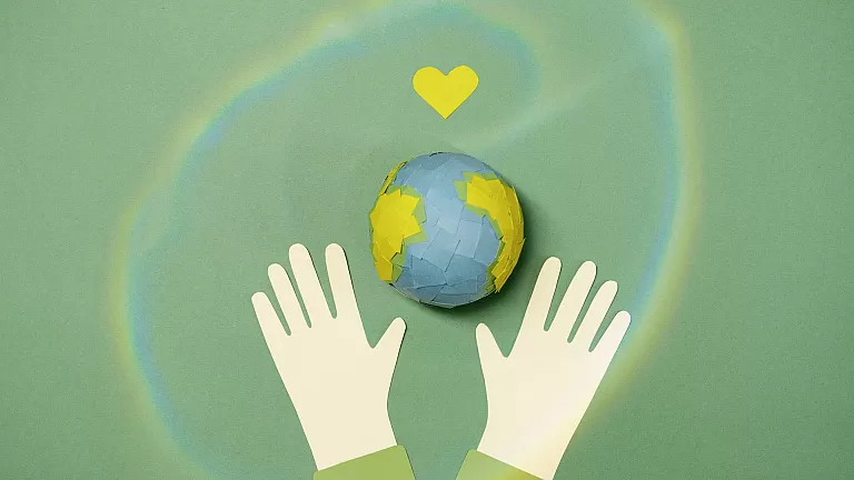 A paper-crafted planet earth, held by two hands with a green heart over it.